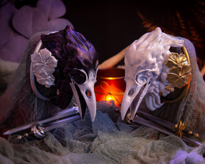 Two resin hair bun covers set up on stands, with their resin sword pins. The bun covers are ravens with gilded geraniums.